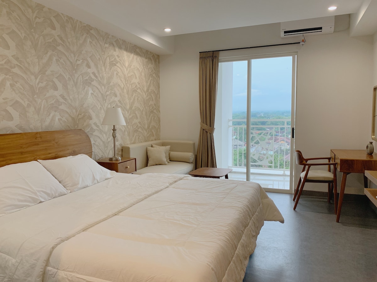 A spacious bedroom features a queen size bed with a light-colored duvet. A small sofa is placed near a large window, offering a view of the surrounding area. A wooden desk and chair are positioned near the entrance, with soft lighting enhancing the room's ambiance.