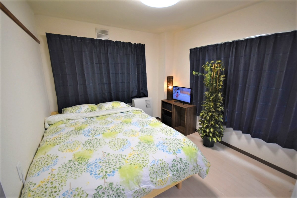 A cozy bedroom features a spacious bed adorned with a floral-patterned duvet. Dark curtains frame the windows, allowing for privacy. A small flat-screen television is positioned on a shelf, alongside a decorative plant, contributing to a relaxed ambiance.