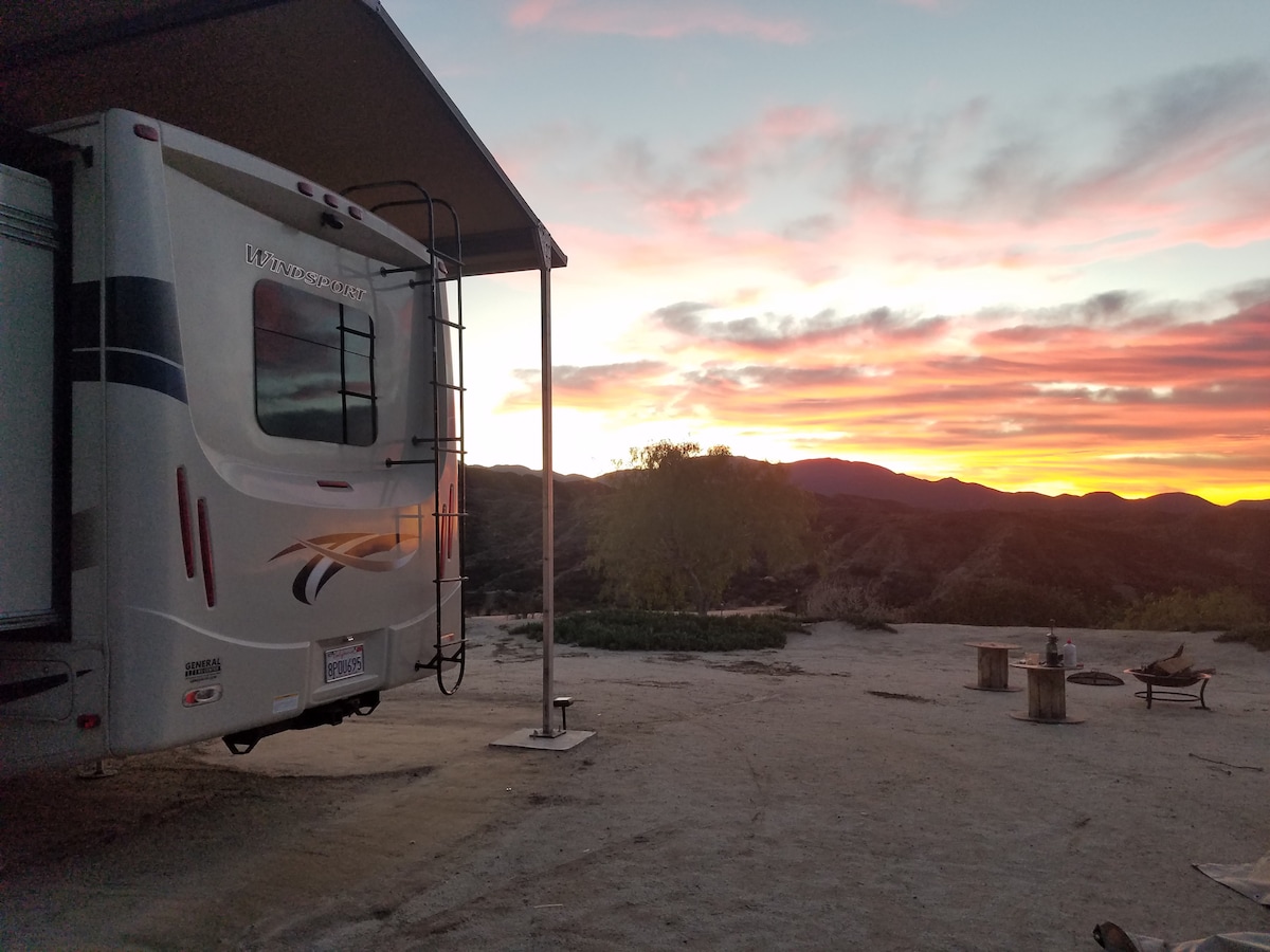 A motorhome is positioned under an awning, framed by a breathtaking sunset that casts warm hues across the sky. The hillside landscape is visible in the background, with tranquil mountains adding depth to the scene. A fire pit and seating area are set nearby.