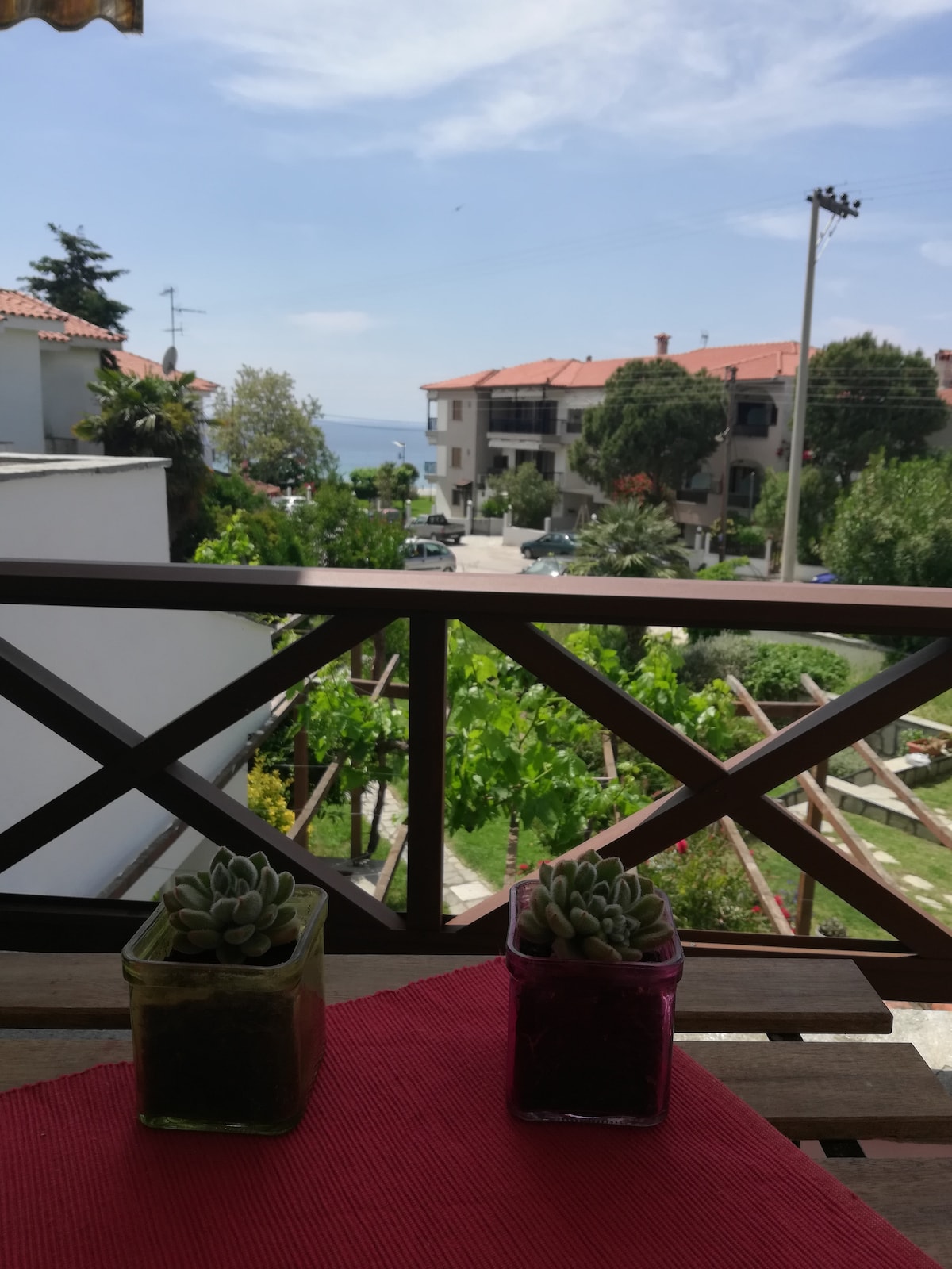 A view from a balcony presents a glimpse of the sea in the distance. Lush greenery and well-maintained gardens are visible below, complemented by vibrant potted succulents on the railing. The charming neighborhood features buildings with terracotta roofs, creating a serene setting.