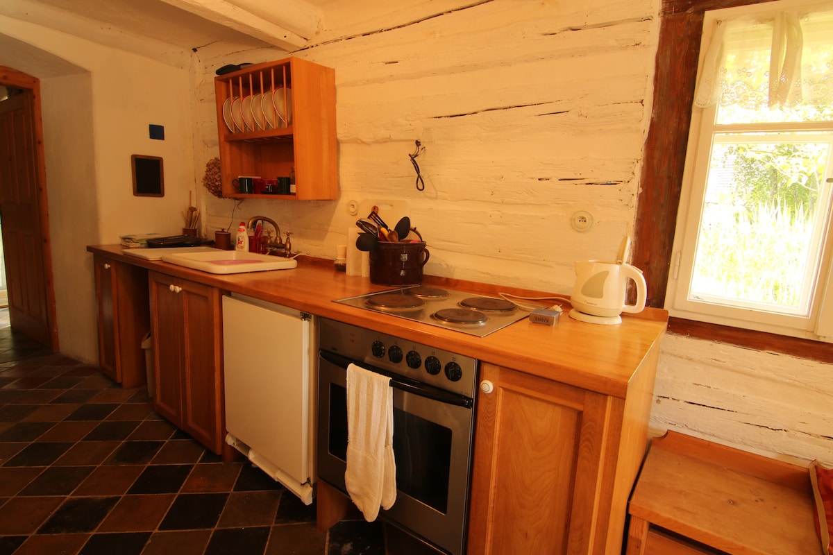 The kitchen features a wooden countertop with essential appliances, including a stove, kettle, and refrigerator. A dish rack is mounted above the counter, while the tiled floor adds texture. Natural light enters through a nearby window, highlighting the rustic charm of the space.