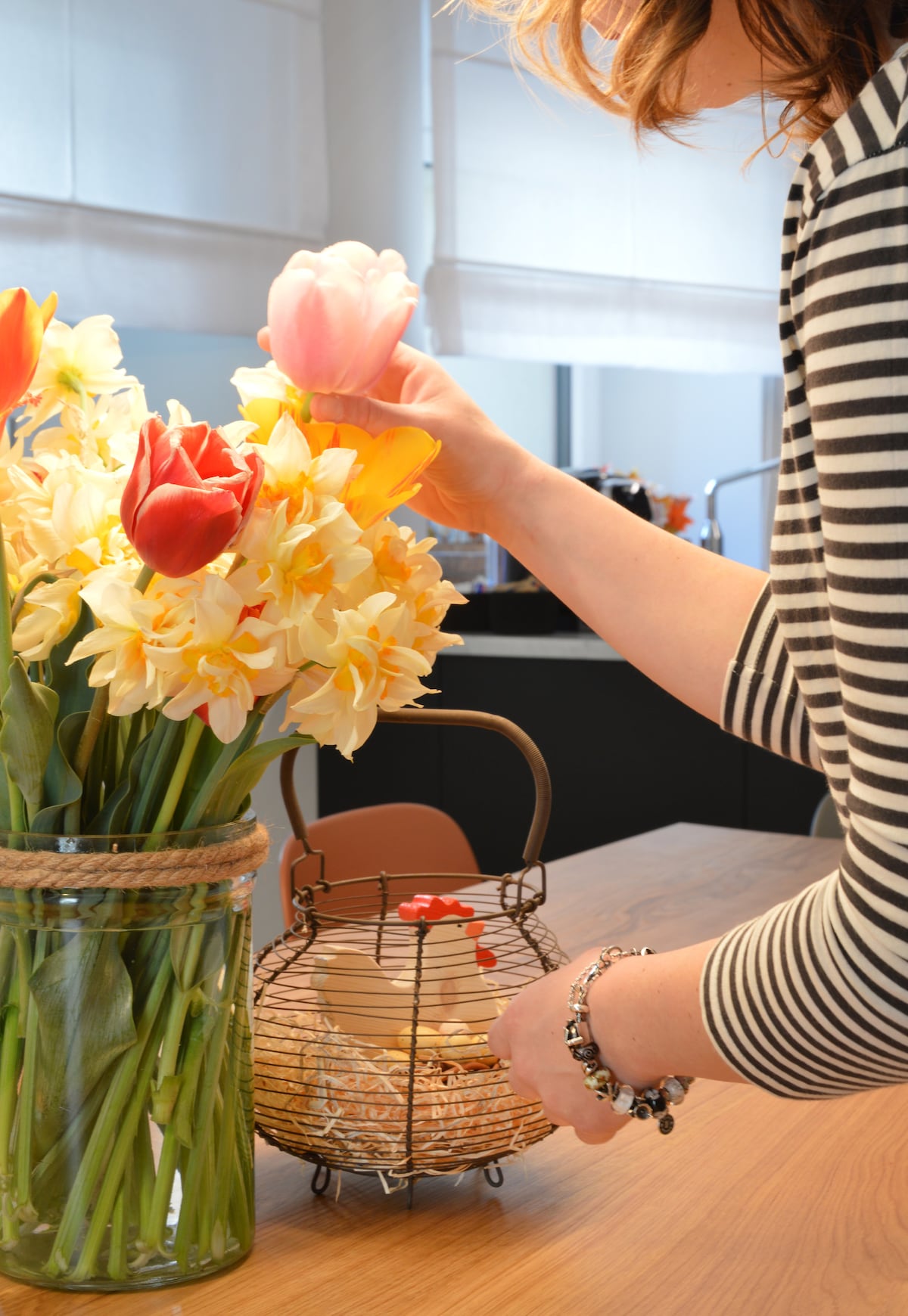 A vibrant arrangement of tulips and daffodils is displayed in a glass vase on a wooden table. A hand is seen adjusting the flowers, while a small wire basket nearby holds assorted items. Natural light is filtering through sheer window treatments.
