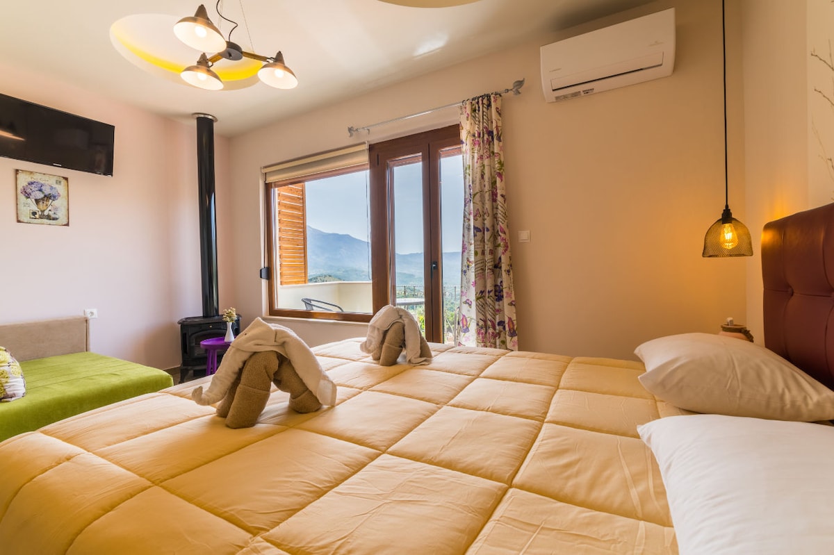 A comfortable guest room is presented with a spacious yellow bedspread and neatly arranged pillows. A view of the mountainous landscape is visible through the large window, complemented by soft curtains. An air conditioning unit and a flat-screen TV are mounted on the wall for convenience.