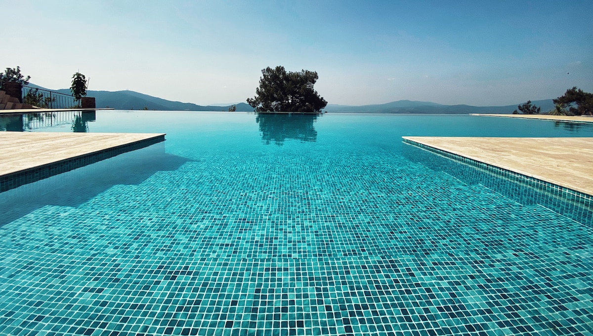 An infinity pool stretches into the horizon, its clear blue water reflecting the sky. Surrounding the pool are sun-drenched stone patios, and distant mountains provide a serene backdrop, enhancing the tranquil atmosphere of the retreat.