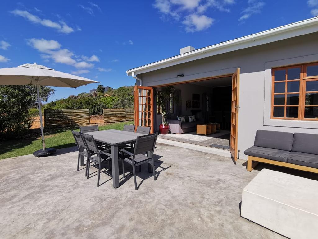 A patio area is featured with a large table and six chairs, providing a space for outdoor dining. An umbrella offers shade, and a comfortable sofa arrangement is positioned nearby. The cottage's door opens to the inviting indoor space, surrounded by natural greenery.