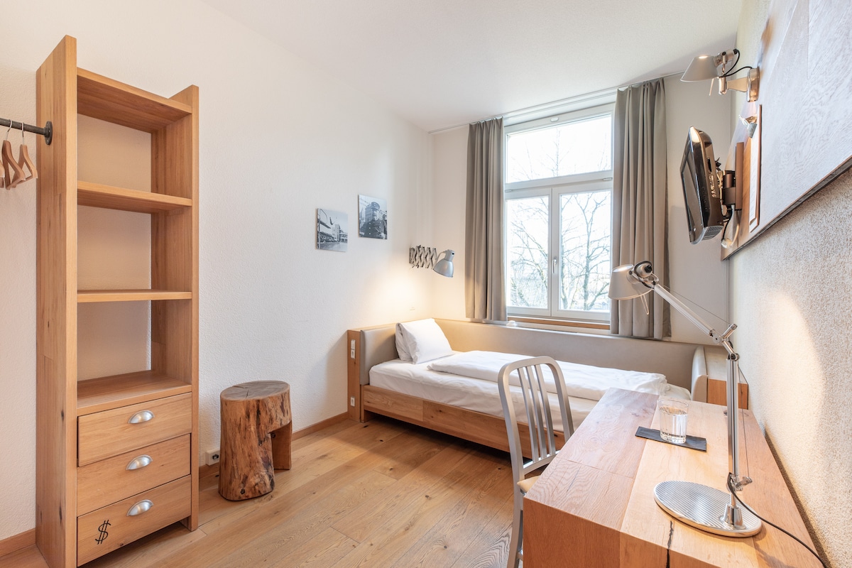 A simple room is shown with light wooden furniture. A spacious bed is positioned against the wall under a window, allowing natural light to enter. A wooden shelving unit with three drawers is present, along with a desk featuring a lamp and a small glass of water.