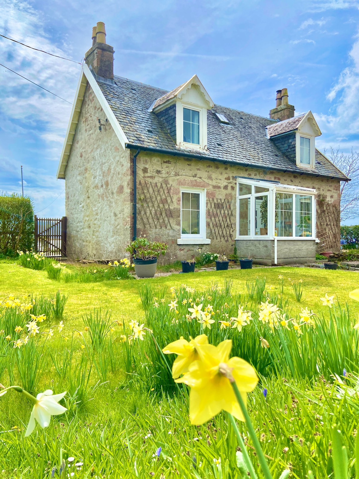 A charming stone cottage is surrounded by a vibrant garden filled with blooming flowers. The house features a gabled roof and large windows that invite natural light. A neatly manicured lawn is visible in the foreground, with a variety of plants adding color to the scene.