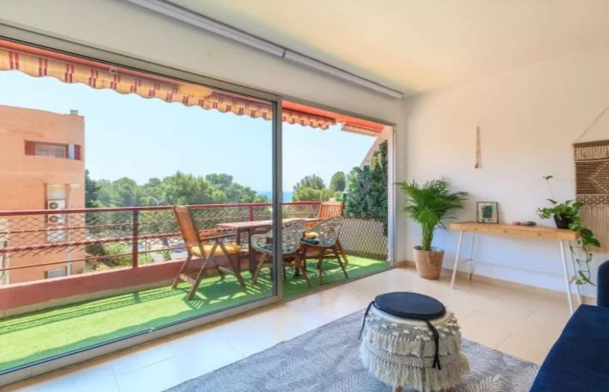 A bright living area features large glass doors that open to a private balcony, showcasing outdoor seating with two chairs and a small table. Lush greenery is visible outside, enhancing the serene atmosphere. A small potted plant and a decorative wall hanging add warmth to the interior.