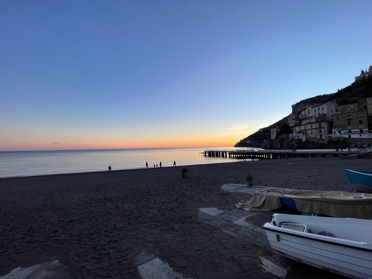 A serene beach scene captures the calm waters reflecting the colors of dusk. Silhouettes of people are seen along the shoreline, while boats are moored in the foreground. The coastline features gentle slopes and buildings rising on the hill in the distance.