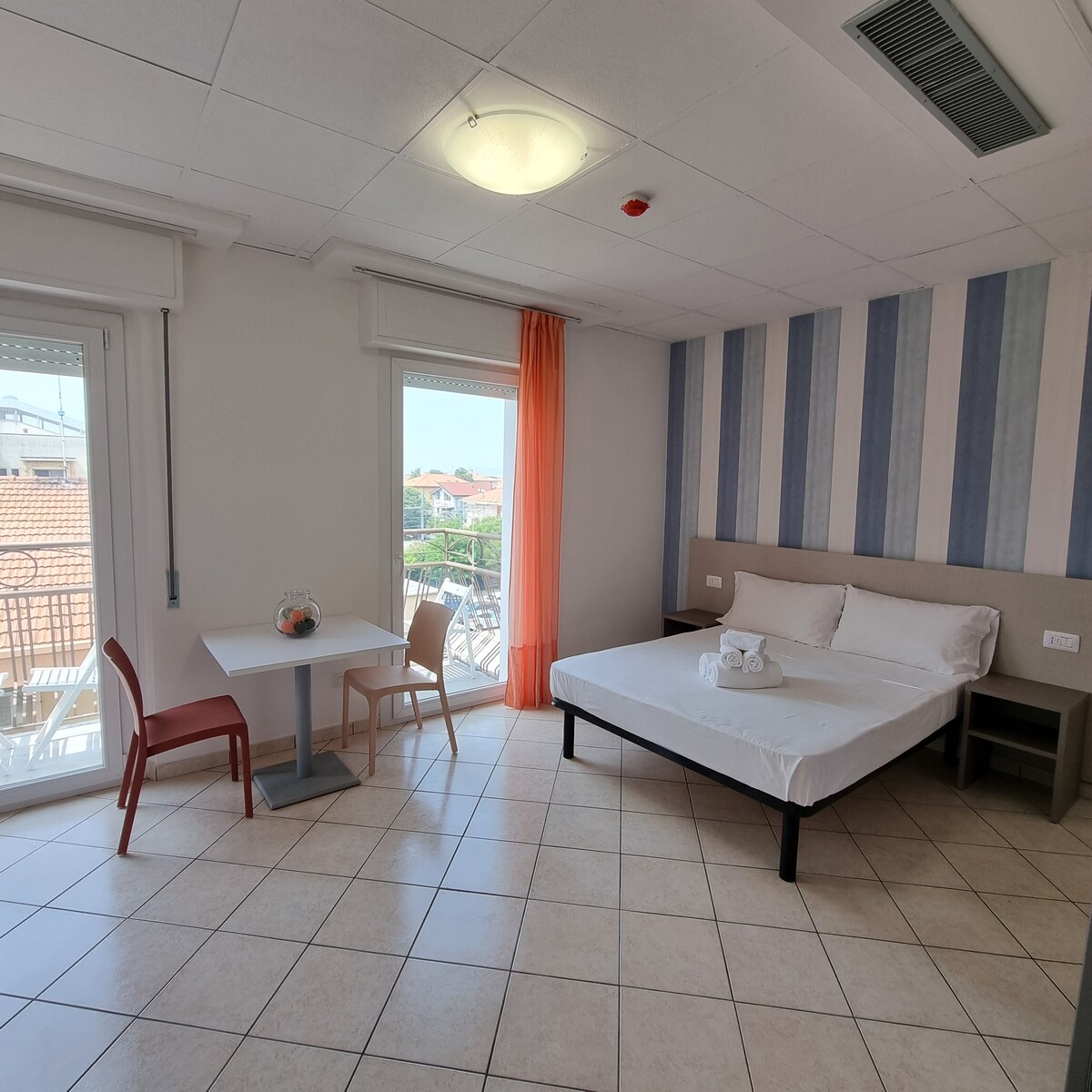 A bright bedroom features a spacious bed and a small table with chairs. Large windows allow natural light to fill the room, with orange curtains adding a touch of color. The tiled floor enhances the open and airy feel of the space.