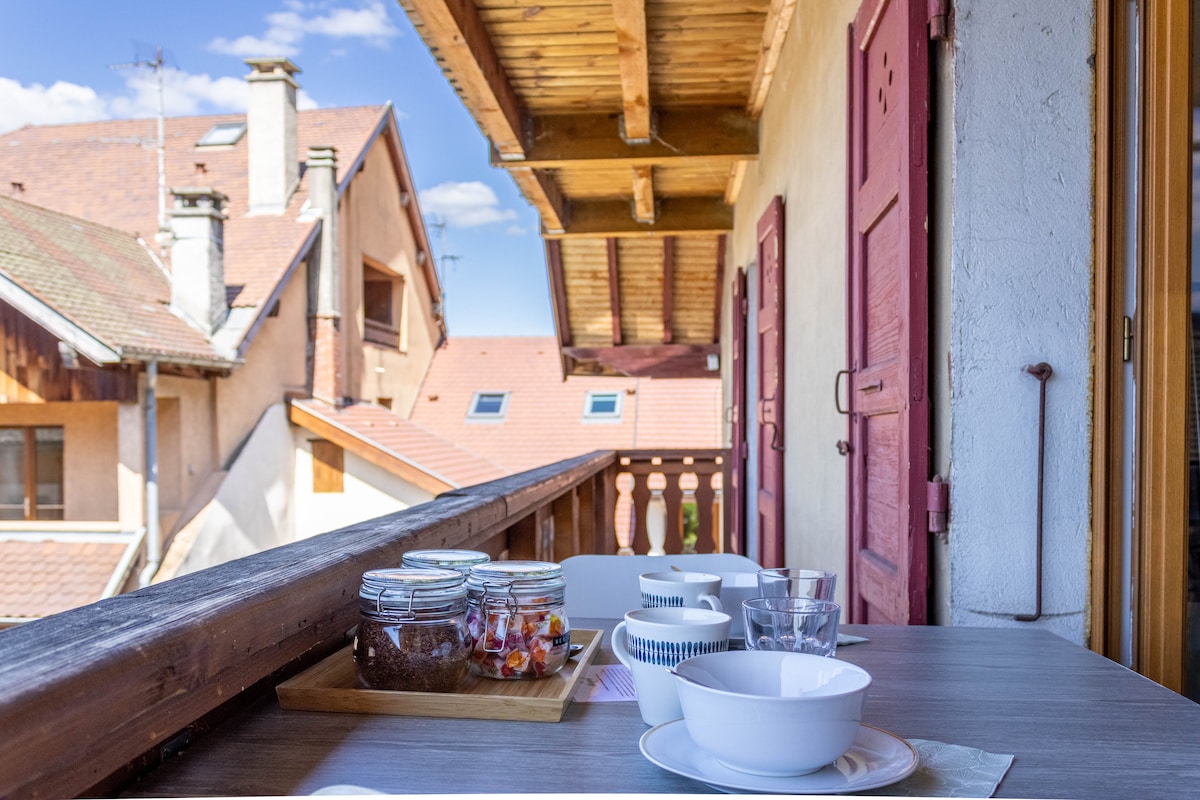 A wooden balcony with a railing overlooks neighboring rooftops. A simple table is set with bowls, glasses, and a tray featuring jars. Soft natural light fills the space, highlighting the ceiling beams and providing a relaxed ambiance.