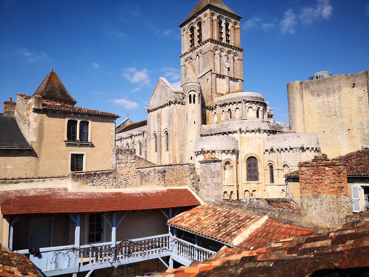 A historic church tower rises prominently against a blue sky, surrounded by traditional stone buildings. The rooftops below feature terracotta tiles, with a wooden balcony adding architectural charm. The scene captures the essence of the medieval architecture prevalent in Chauvigny.