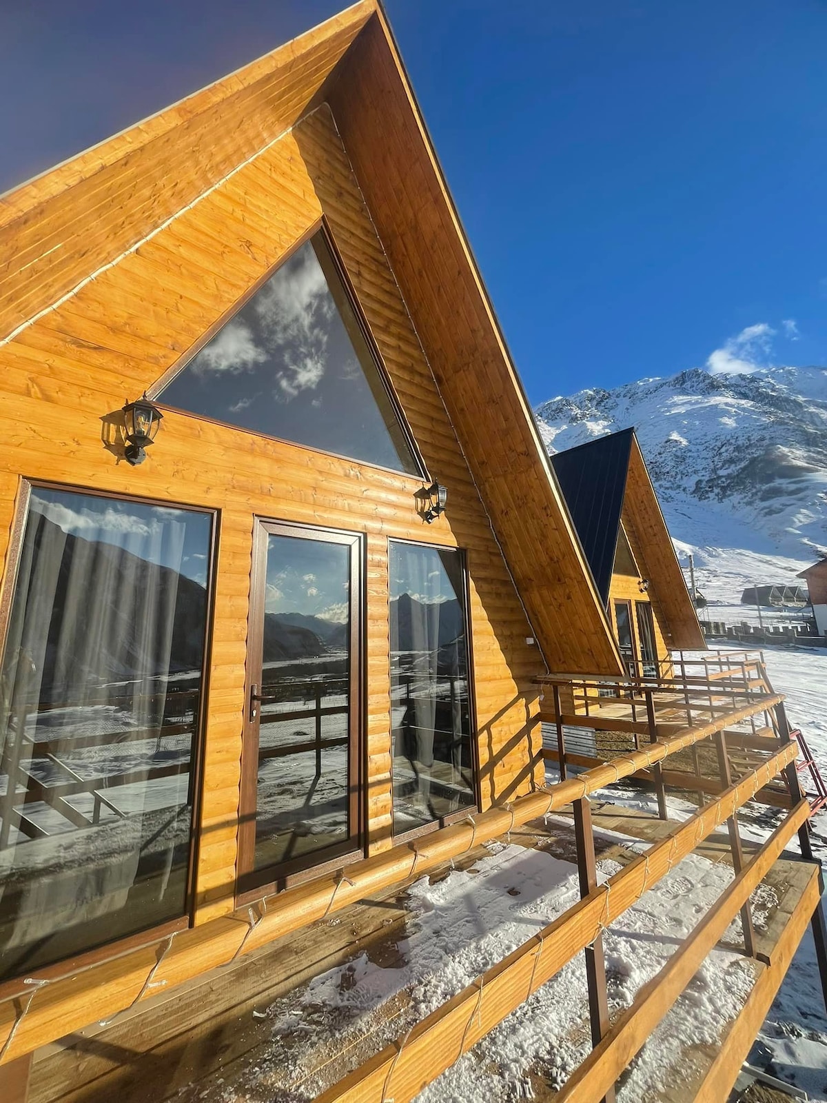 A cabin with a triangular wooden design is shown, featuring large glass doors that provide access to a balcony. Snow covers the ground outside, and mountains are visible in the background under a clear blue sky. Natural light highlights the wooden textures.