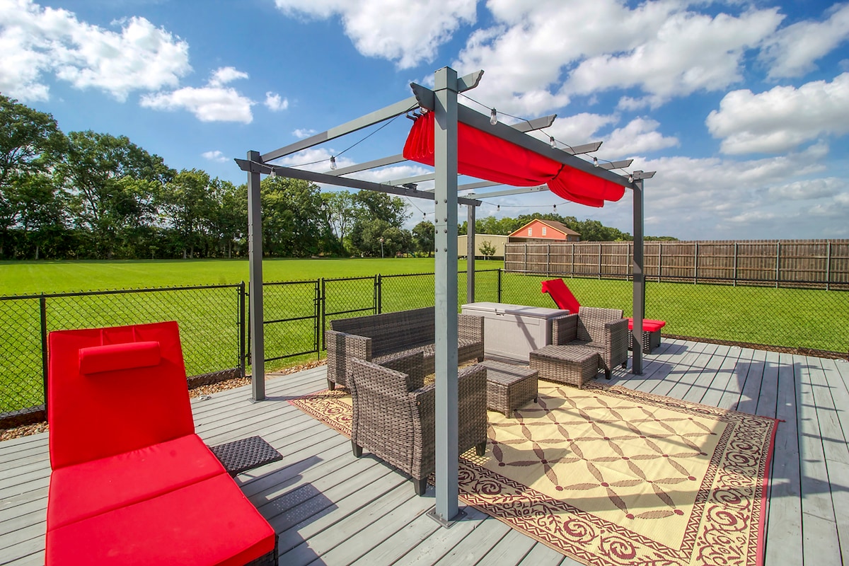 A spacious outdoor deck is furnished with stylish seating, including a set of wicker sofas and lounge chairs in vibrant red. A large, patterned rug anchors the seating area under a shaded pergola, with a green lawn extending beyond a privacy fence in the background.