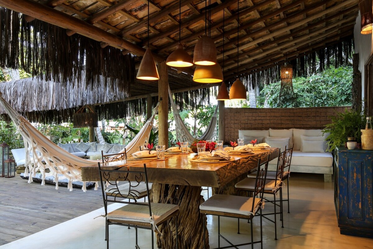 An outdoor dining area is featured, highlighting a large wooden table surrounded by chairs. Soft lighting is provided by hanging lamps, creating a relaxed ambiance. Hammocks are positioned nearby, offering a comfortable space for relaxation amidst lush greenery.