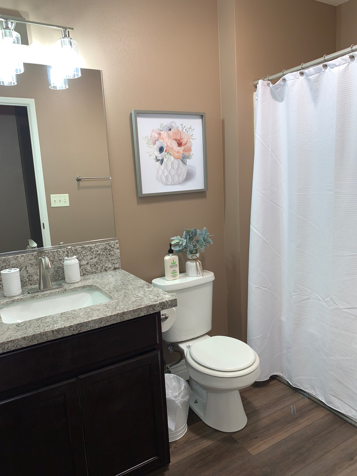 A well-lit bathroom features a granite countertop with a sink and modern fixtures. A mirror above the sink reflects soft lighting from three overhead bulbs. A white shower curtain separates the bathing area, and a floral art piece adds a touch of color to the wall.