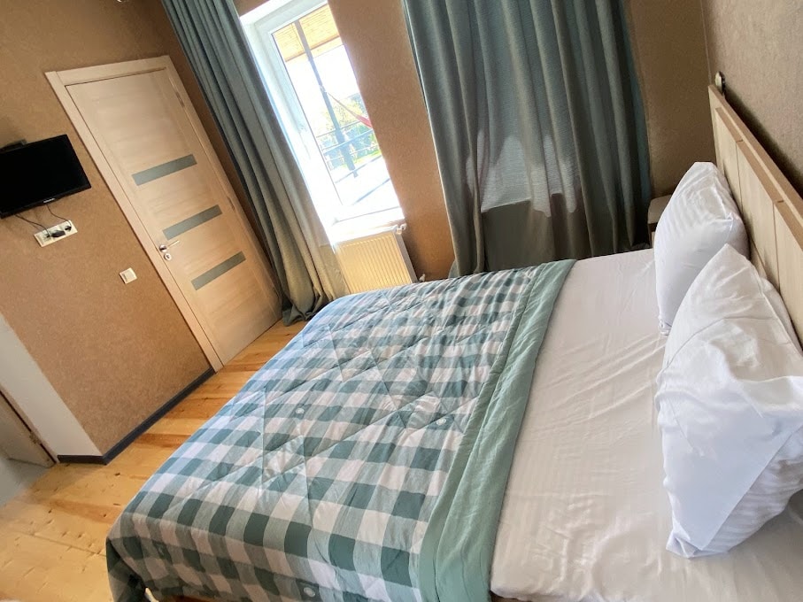 A well-appointed sleeping area features a spacious bed with a light-colored checkered blanket. The bed is positioned near a window with beige curtains, allowing natural light to brighten the room. A wall-mounted television is visible, and a wooden door leads to the hallway.