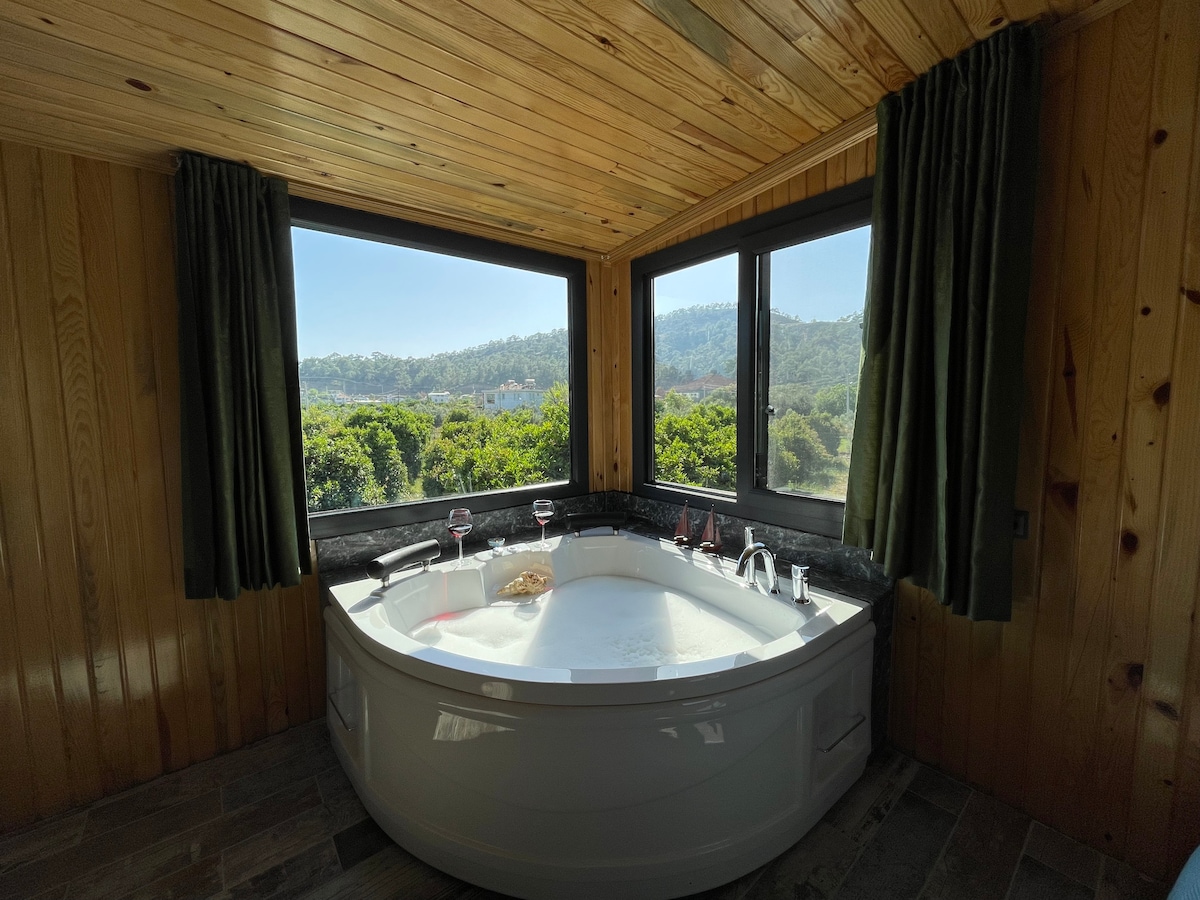 A spacious jacuzzi is positioned by large windows, offering panoramic views of the surrounding greenery. Natural light floods the area, complementing the wooden ceiling and soft green curtains that frame the scene. Glasses are set nearby, enhancing the inviting atmosphere of relaxation.