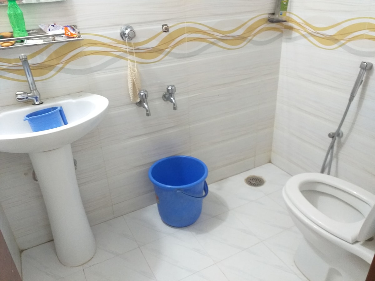 The bathroom features a white pedestal sink and a modern toilet with a flush system. A blue bucket is placed on the tiled floor, and two water taps are visible on the wall. A shelf above the sink holds various toiletries.
