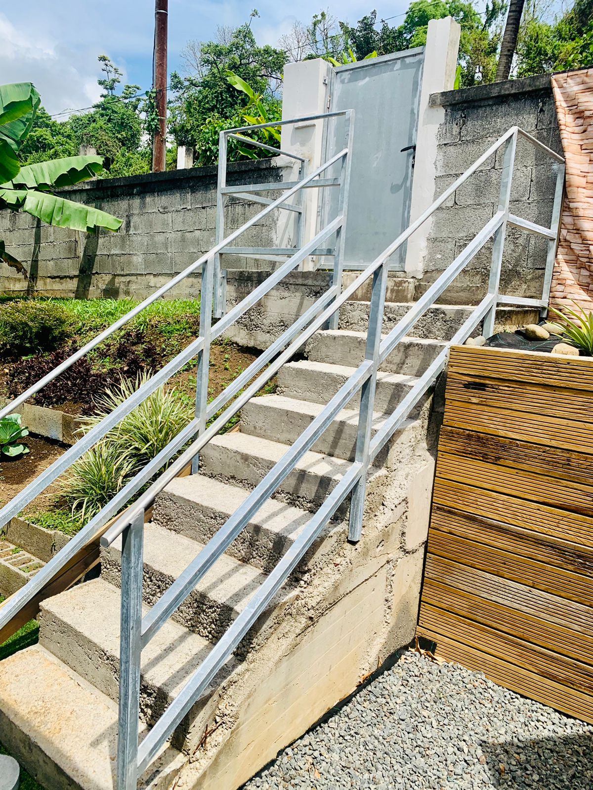 A set of sturdy metal stairs is shown, leading up from a landscaped outdoor area to a gated entrance. The stairs are bordered with railings for safety, and greenery is visible along the side, contributing to a serene environment.