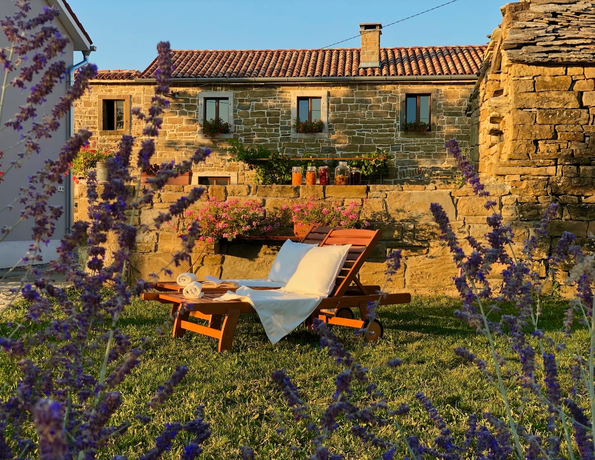 A wooden lounge chair is positioned on a grassy area, surrounded by blooming lavender. In the background, a stone building with window boxes filled with flowers is visible, offering a serene outdoor space for relaxation.