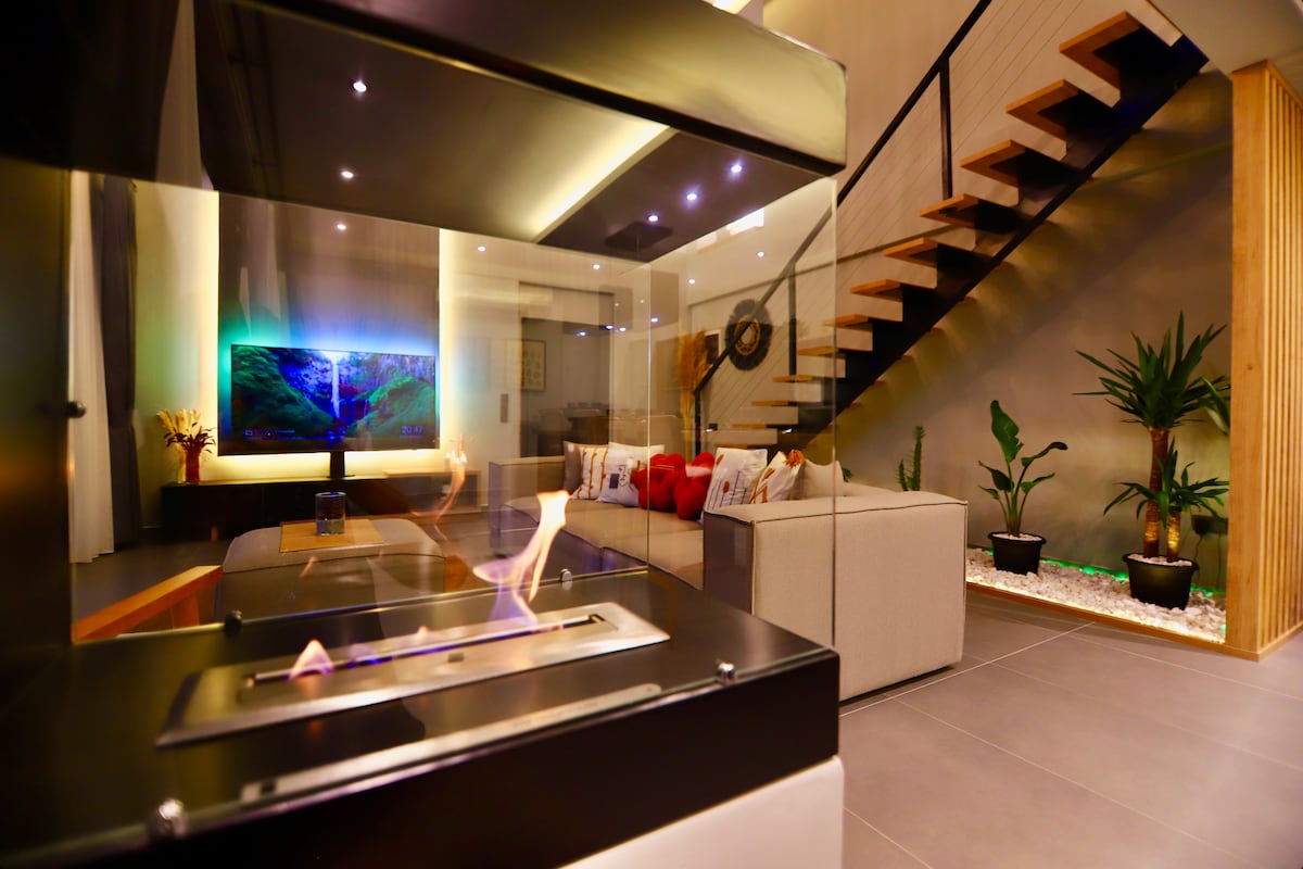 A modern living area features a glass-enclosed fireplace that offers warmth and ambiance. A large TV is mounted above a sleek console, while a spacious sofa adorned with colorful cushions invites relaxation. Natural plants add a touch of greenery to the minimalist design.