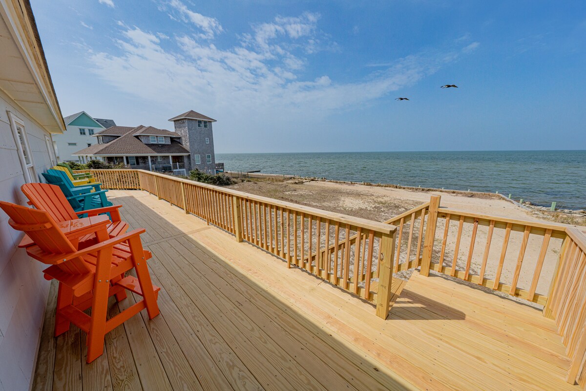 A wooden deck features colorful adirondack chairs facing the water. The shore is visible in the distance, with gentle waves lapping at the sand. A clear sky is overhead, and two birds can be seen flying across the serene landscape.