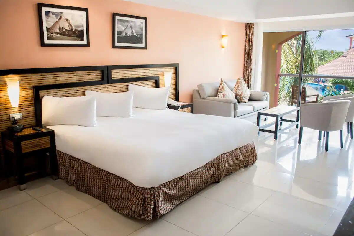 A spacious bedroom features a large bed with crisp white linens, complemented by decorative accent pillows. A cozy seating area includes a light sofa and a side table, with views through large glass doors that open to a balcony overlooking the resort landscape.