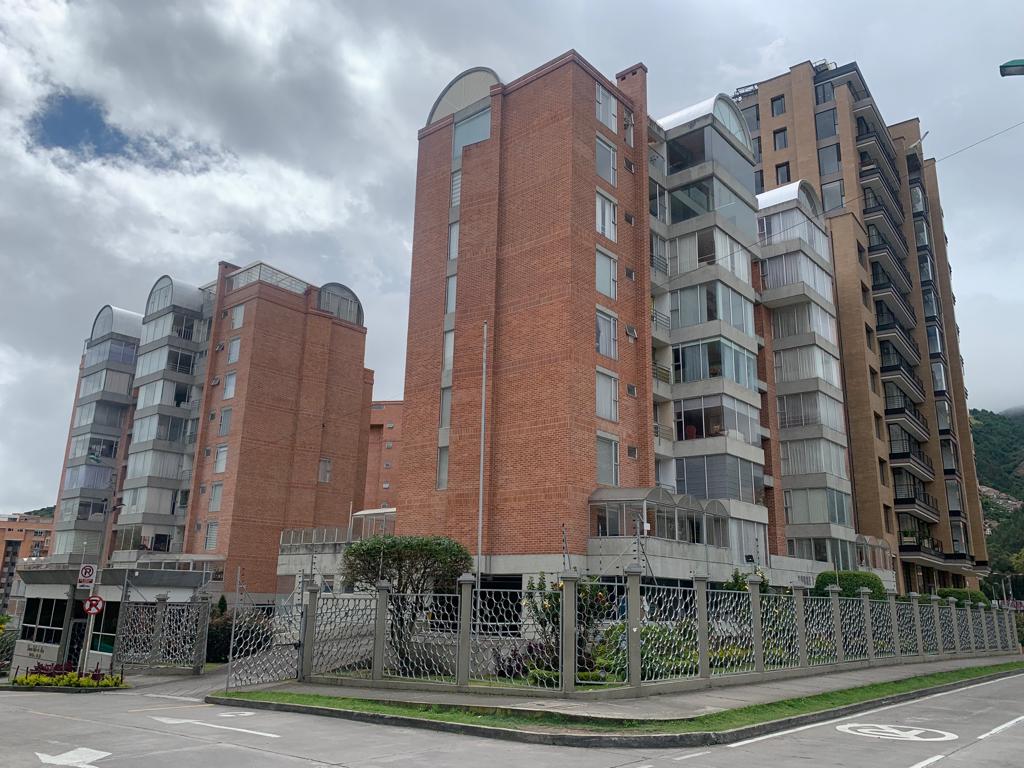 The image showcases a modern, multi-story building comprised of red brick and glass elements. The structure features several balconies and a landscaped area with greenery in the foreground. A clear sky and distant hills are visible in the background.