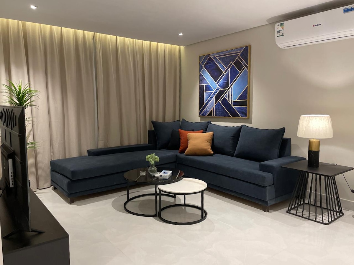 A living area features a dark blue sectional couch adorned with orange and white pillows. A circular coffee table is positioned between two side tables, one with a lamp. Soft lighting highlights the beige curtains and a geometric art piece on the wall.