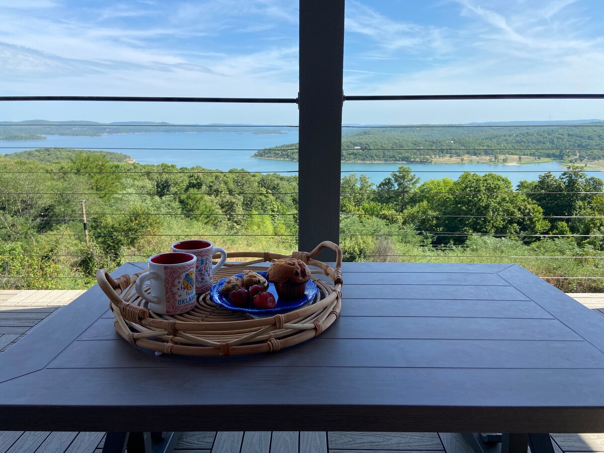 A patio table is set with a tray holding coffee mugs, a plate of strawberries, and baked goods. The sweeping view of the tranquil lake and surrounding greenery is visible in the background, enhancing the outdoor dining experience.