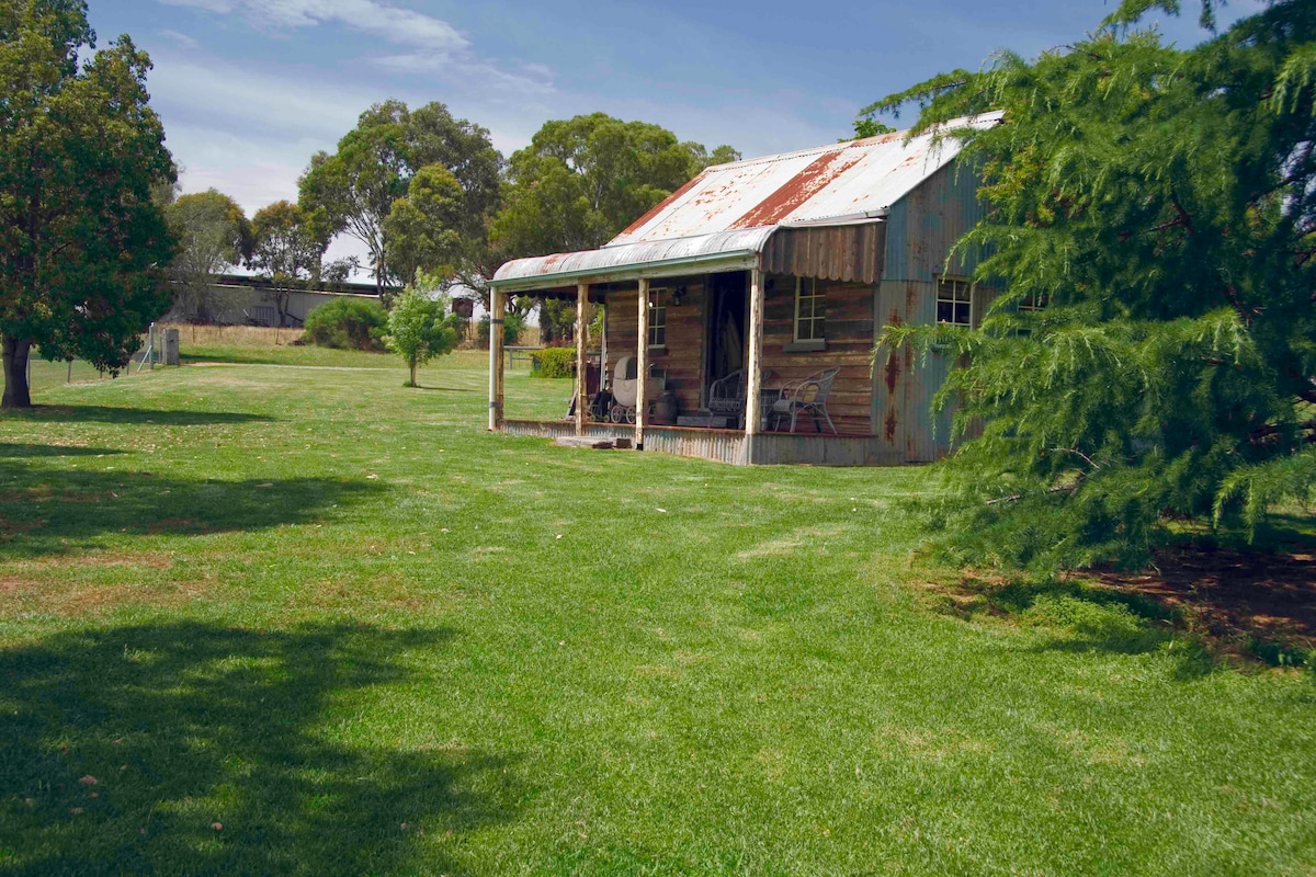 A traditional replica cottage features a rusted corrugated metal roof and a wooden veranda. The outdoor area is expansive, set in a grassy landscape with trees providing shade. The home is framed by patches of greenery, creating a peaceful rural setting.