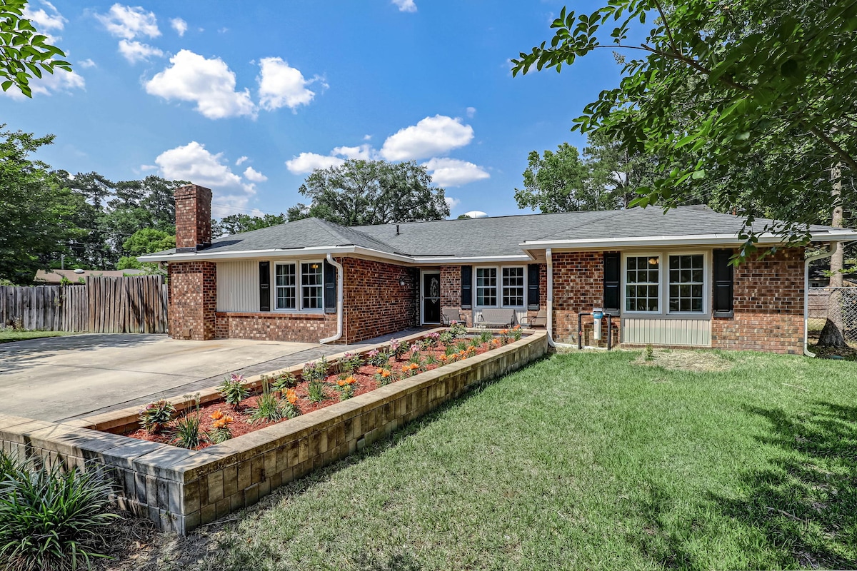 The exterior of a single-story brick house is displayed, featuring a wide driveway and a landscaped garden with flower beds. Green grass surrounds the home, and tall trees provide shade in the background. A fenced area is visible, enhancing privacy.