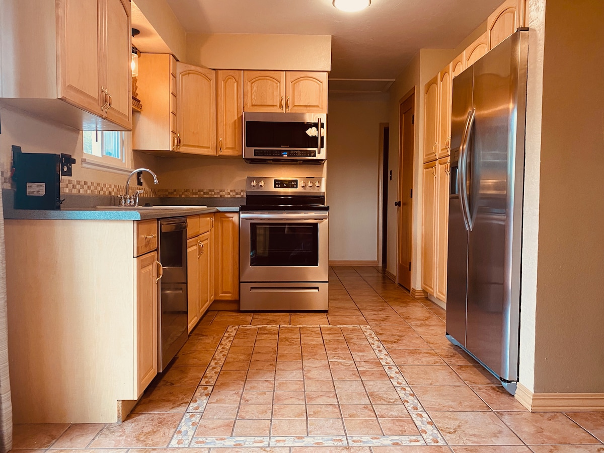 A well-equipped kitchen features light wood cabinetry and stainless steel appliances. A built-in microwave and oven are positioned above a smooth cooktop. The spacious countertop is complemented by a tiled floor, providing a practical and inviting workspace.