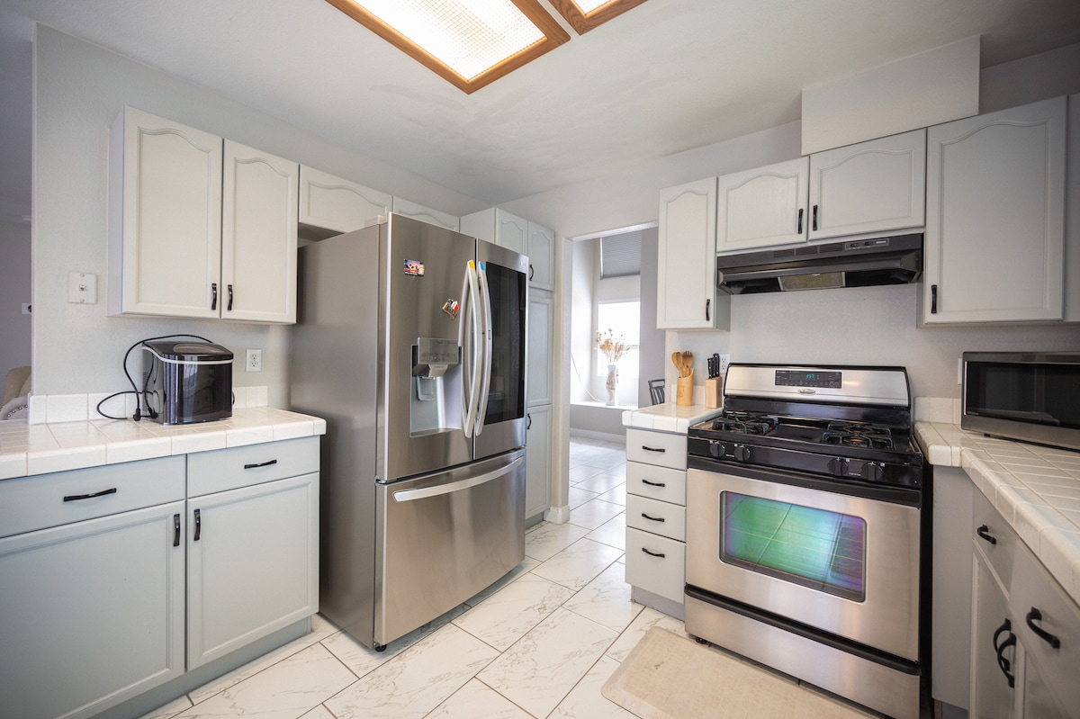 The kitchen features modern appliances including a stainless steel refrigerator and a gas stove. Light-colored cabinets offer storage, while ample counter space provides room for meal preparation. Natural light enters through a nearby window, illuminating the tiled floor.