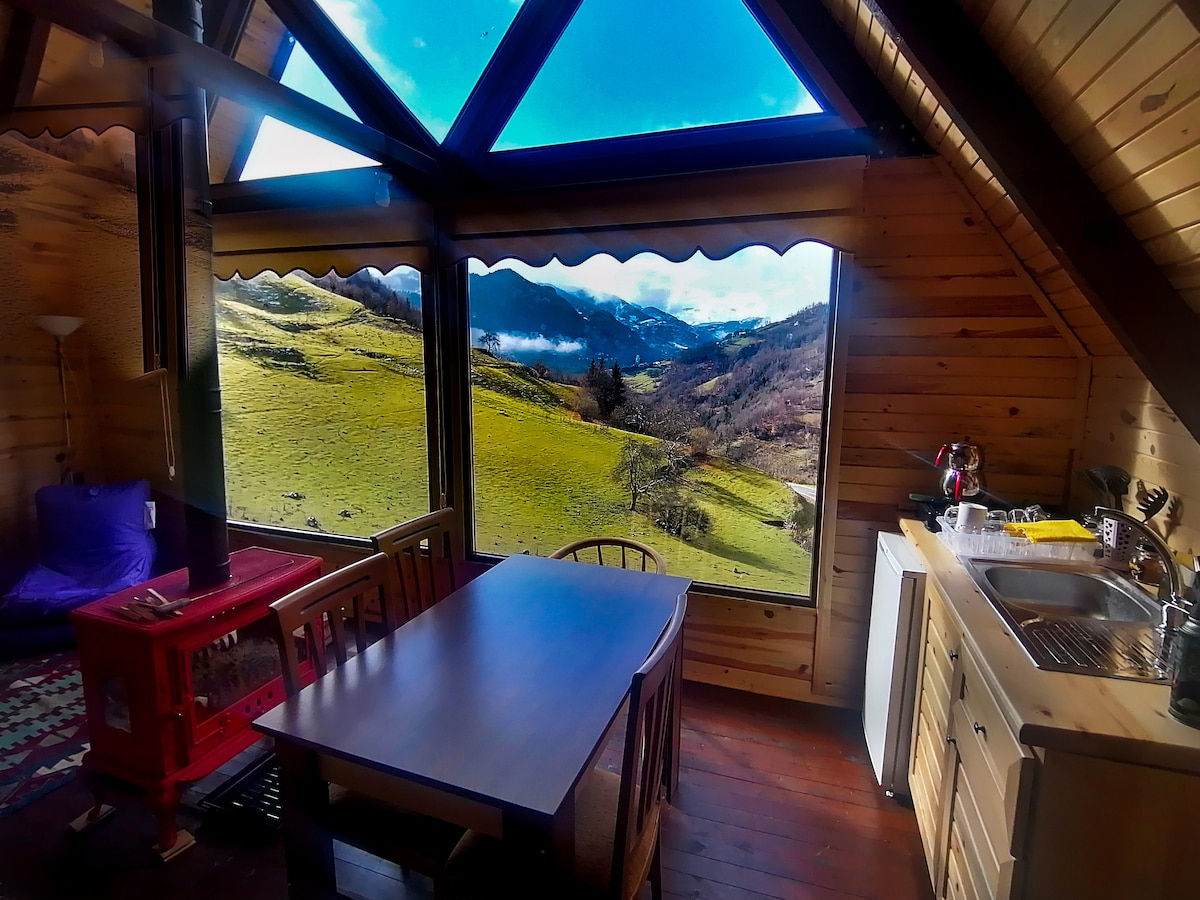 A bright interior space features a wooden dining table surrounded by chairs. Large windows offer sweeping views of green hills and distant mountains. A cozy seating area is positioned near a red stove, while a functional kitchenette is visible in the corner.