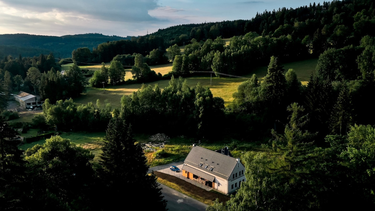 A spacious villa is nestled amidst lush green landscapes, surrounded by trees and open fields. The building features a pitched roof and large windows that reflect the natural surroundings. A quiet road runs nearby, enhancing the sense of tranquility offered by the area.