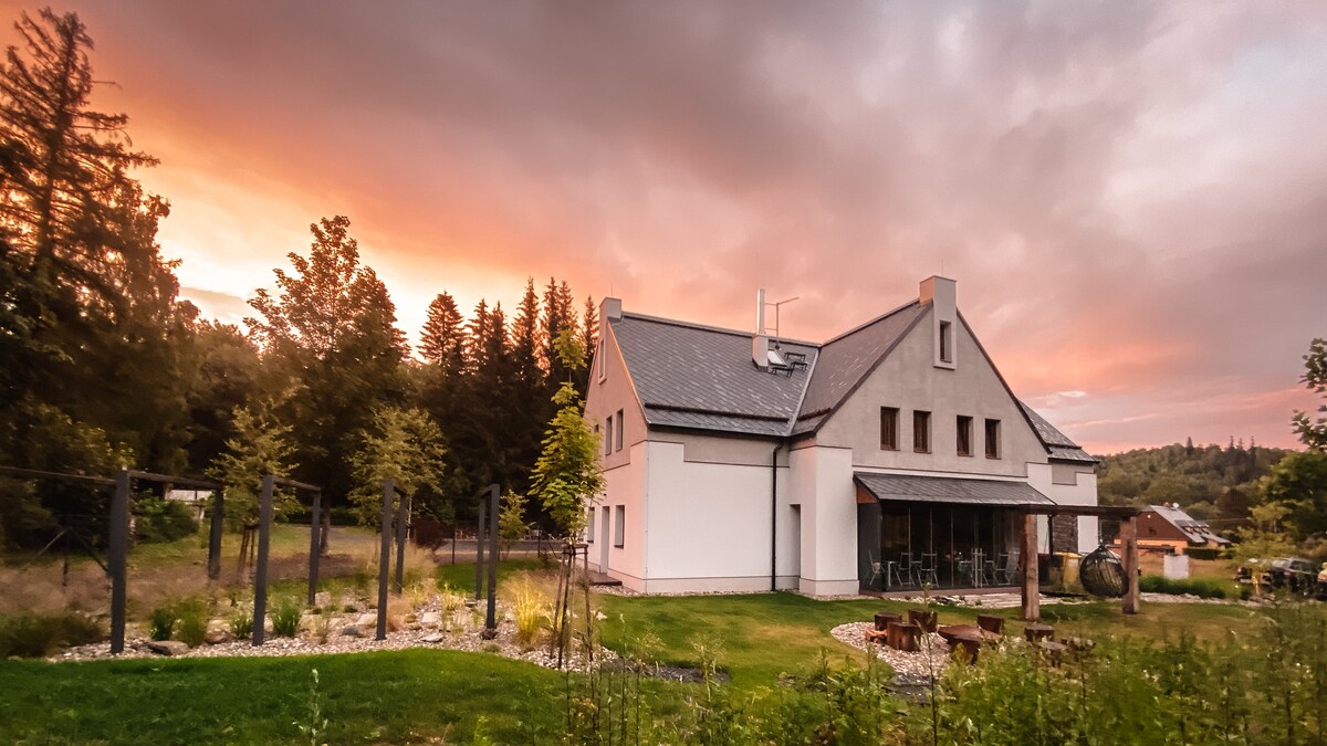 A modern villa is set against a colorful sunset, surrounded by lush greenery. The structure features a combination of stone and plaster, with large glass doors providing views of the tranquil outdoor space and nearby forest.