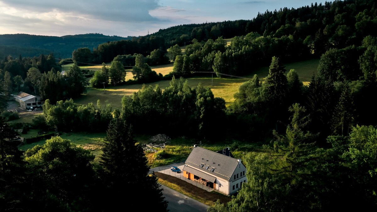 An aerial view of Vila Rasola is presented, highlighting its spacious exterior surrounded by lush greenery. The building is nestled against rolling hills, with fields stretching out in the background. A winding road can be seen, leading towards the entrance of the villa.