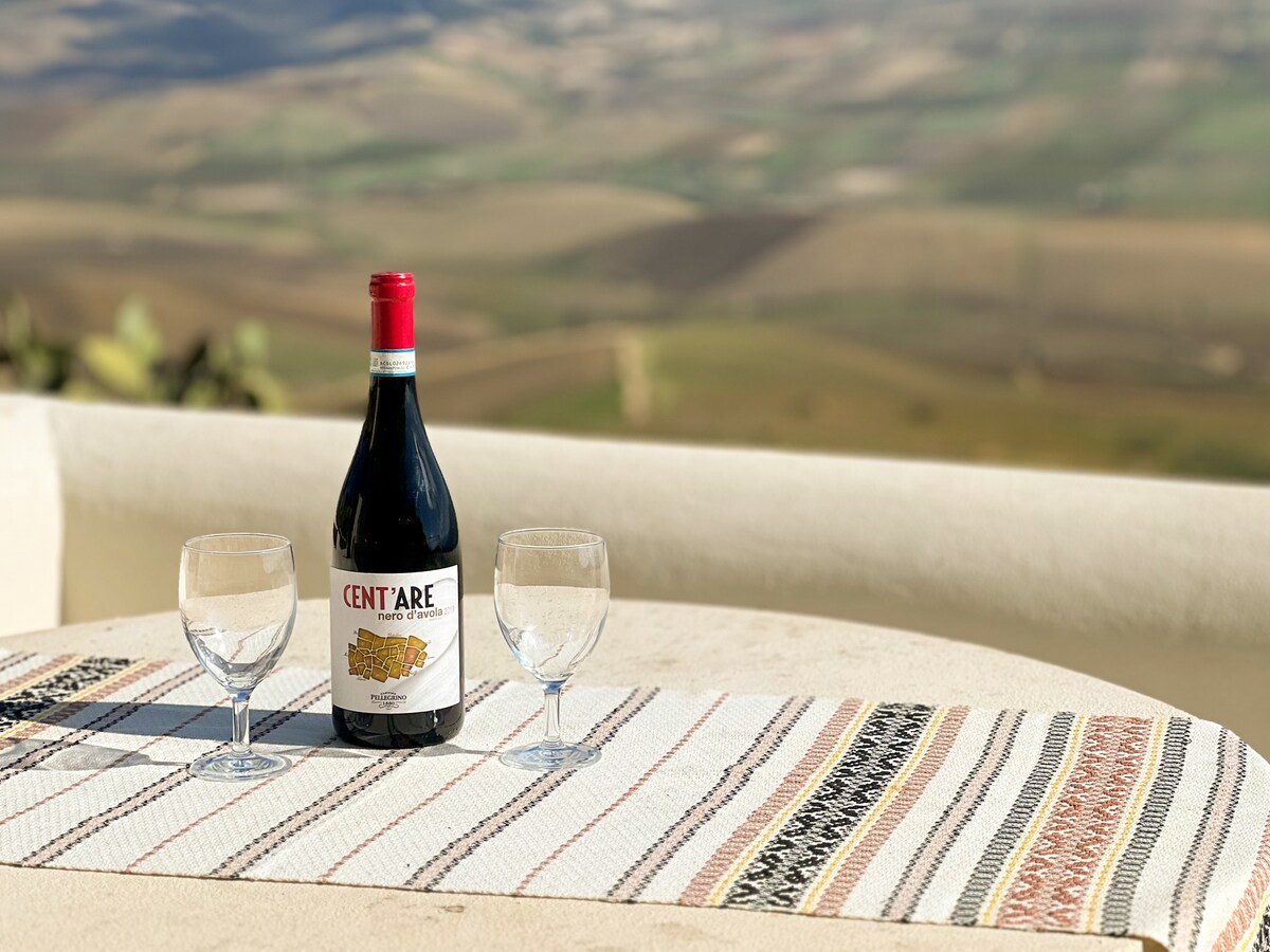 A bottle of wine is placed on a woven table runner, accompanied by two empty glasses. The setting features a round table, with a scenic valley backdrop visible in the distance, showcasing rolling hills and a serene landscape.