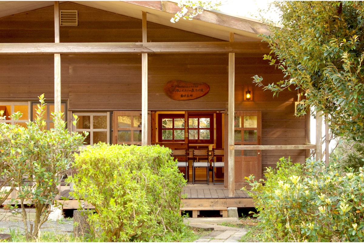 A wooden cottage features a spacious front porch with a rustic sign above the entrance. Large windows are adorned with subtle drapes, revealing a warm interior. Surrounding greenery adds a touch of freshness, creating an inviting atmosphere for relaxation.