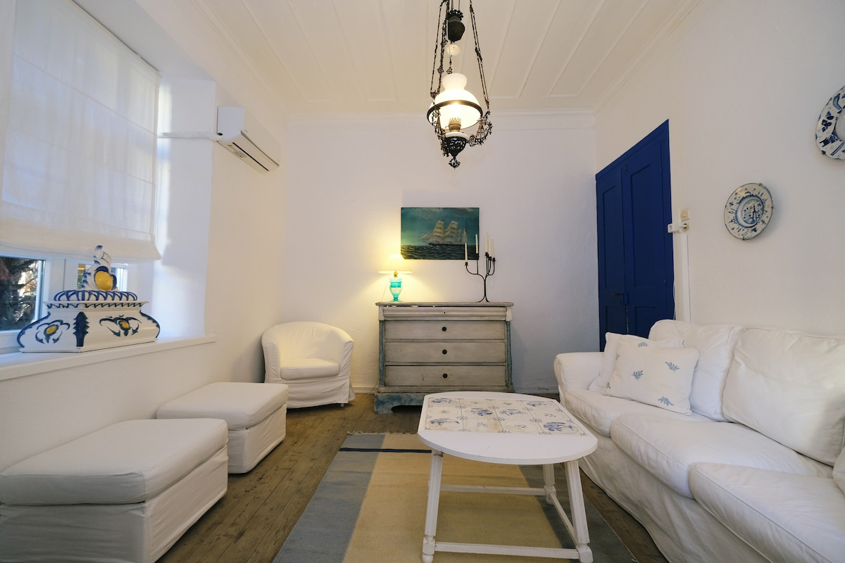 A bright living area features a white sofa set and two armchairs, arranged around a low coffee table. A wooden chest serves as a storage piece, complemented by nautical-inspired artwork on the wall. Natural light filters through a window with light curtains, enhancing the room's airy feel.