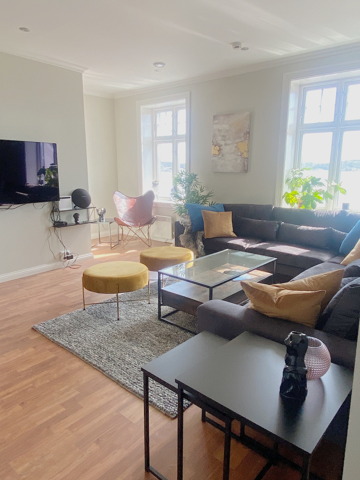 A bright living area features a dark sectional sofa paired with colorful accent pillows. A glass coffee table is centered on a textured area rug. Natural light floods through large windows, illuminating the stylish décor, including a butterfly chair and plants adorning the space.