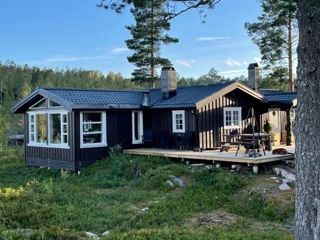 A charming cabin features a dark exterior with large windows reflecting the surrounding nature. A spacious deck is visible, complemented by outdoor seating. The structure is framed by tall trees and a grassy area, creating a serene and natural setting.