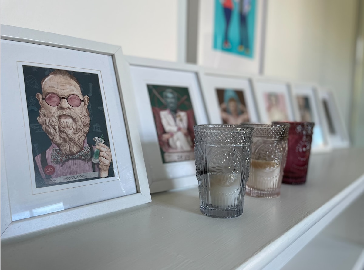 A series of framed art pieces is displayed on a white shelf, complemented by decorative glass candle holders. The holders vary in color, with some containing lit candles, creating a subtle ambiance. The arrangement adds a personal touch to the living area.