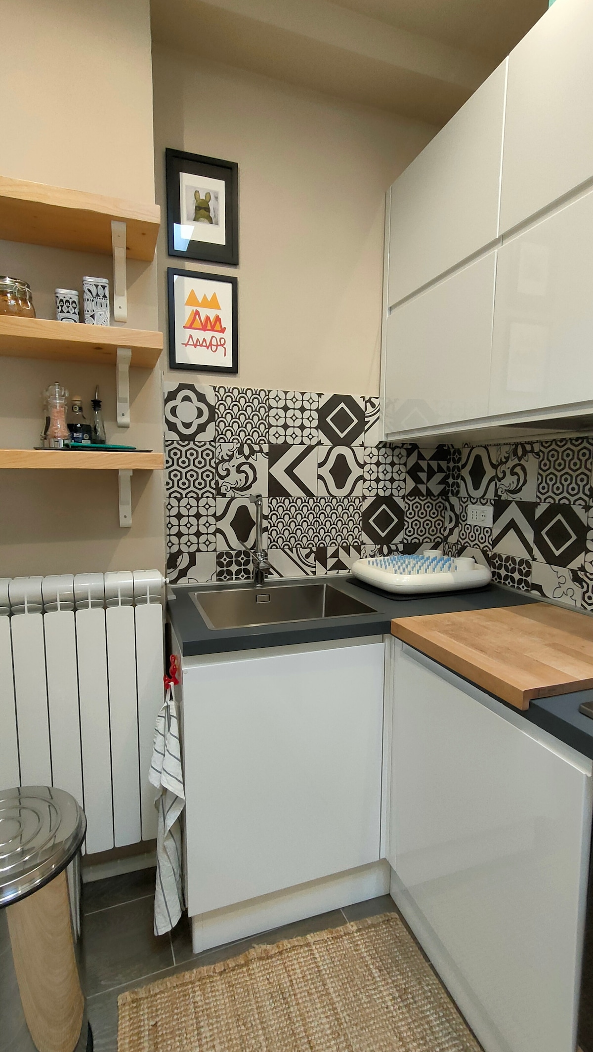 A modern kitchen area features sleek white cabinetry and a decorative black and white tiled backsplash. Open shelves display kitchenware and decorative items. A stainless steel sink is integrated into the countertop, with a small dish rack placed nearby. A woven rug is positioned on the floor.
