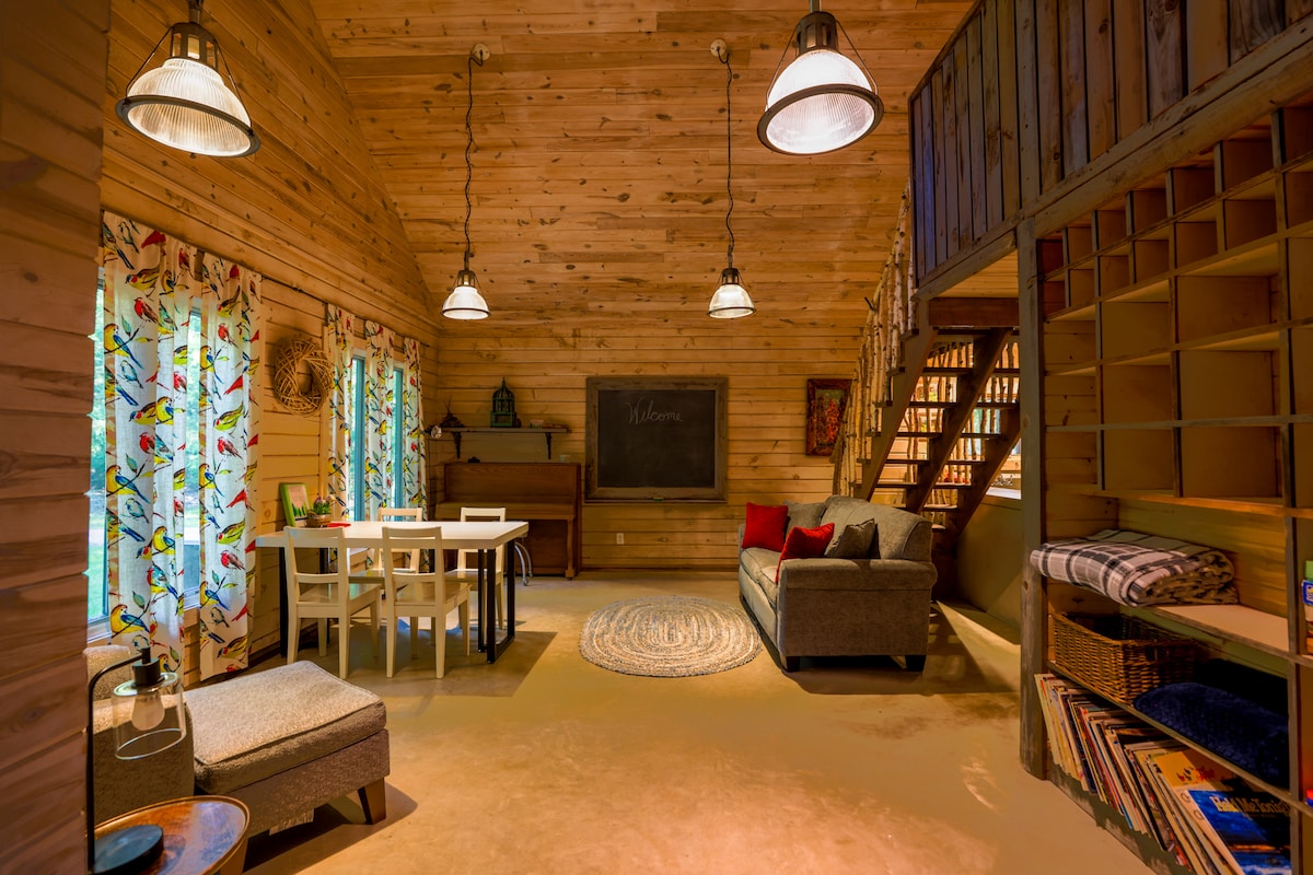 A spacious log cabin interior is highlighted by warm wooden walls and ceiling. Natural light enters through two window pairs adorned with colorful curtains. A dining table and chairs are positioned beside a comfortable seating area featuring a lounge chair and sofa. A staircase leads to the loft.