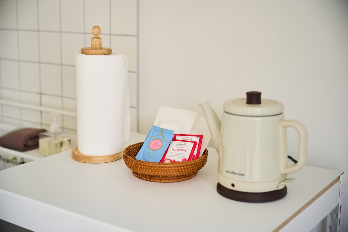 A tidy counter displays a kettle next to a woven basket, which holds various tea bags. A paper towel holder stands nearby, complementing the clean white and muted decor of the kitchen space.