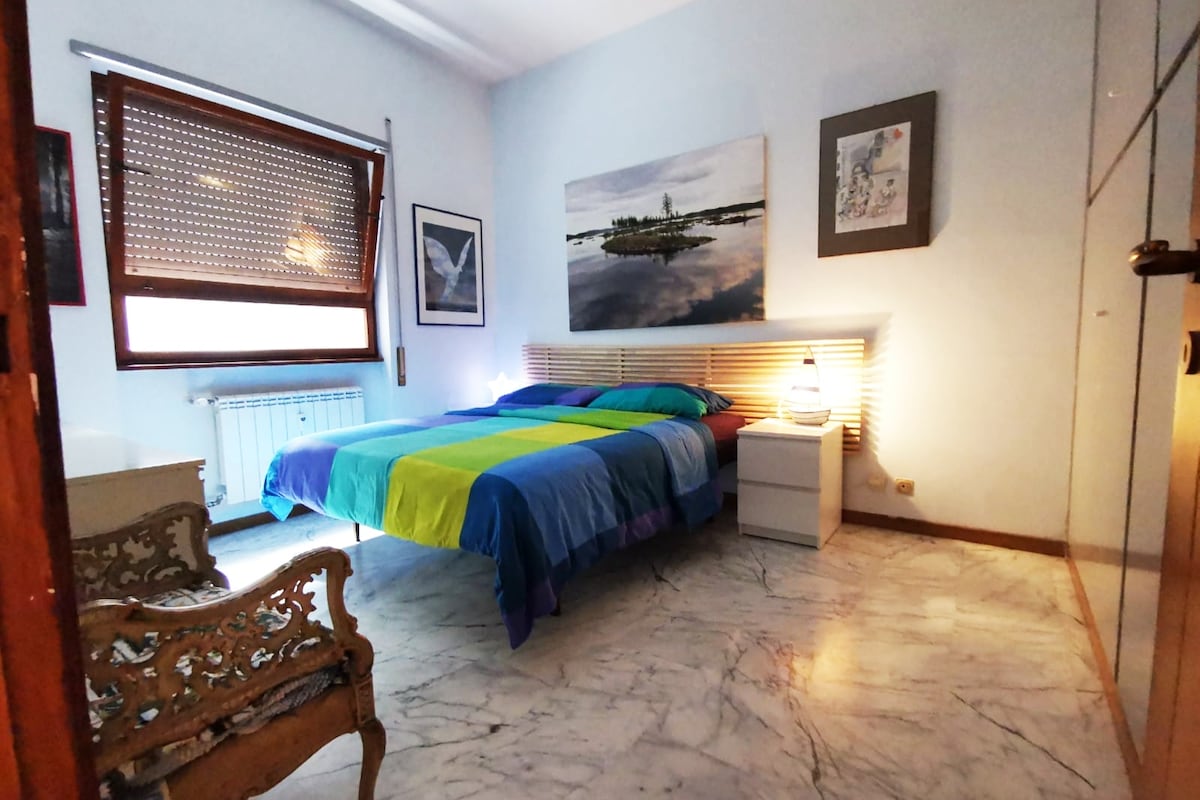 A spacious bedroom features a large bed with a colorful striped blanket. Artwork adorns the walls, and a nightstand with a lamp is positioned beside the bed. Natural light enters through a window, enhancing the room's inviting ambiance.