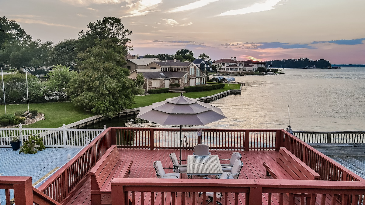 A spacious red deck overlooks a serene waterfront, featuring a table with light-colored chairs and a large umbrella for shade. The calming hues of the sunset reflect on the water, while well-maintained greenery and waterfront properties can be seen in the background.